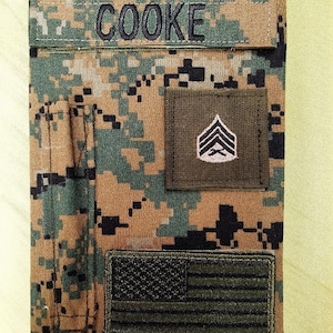 May include: A camouflage pattern notebook cover with the name "COOKE" embroidered in black. It features a sergeant rank patch and an American flag patch. The cover is in shades of green, brown, and black.