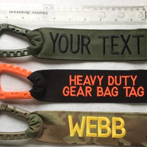 May include: Three bag tags with carabiner clips. One tag is olive green with "YOUR TEXT" in black. Another is black with "HEAVY DUTY GEAR BAG TAG" in orange. The last is camouflage with "WEBB" in yellow.