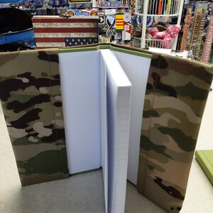 May include: An open notebook with a camouflage cover. The cover features a pattern of green, brown, and tan. The pages are white and lined. The notebook is displayed on a light-colored surface.