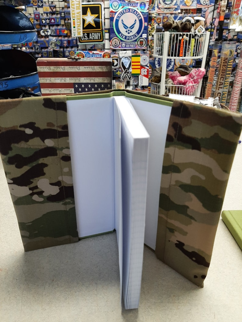May include: An open notebook with a camouflage cover. The cover features a pattern of green, brown, and tan. The pages are white and lined. The notebook is displayed on a light-colored surface.
