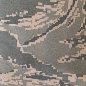May include: Close-up of a fabric with a digital camouflage pattern. The design features a pixelated blend of gray, green, and tan hues, creating a textured appearance. The pattern is a repeating design, suitable for various applications.