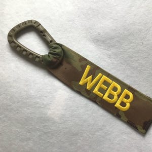 May include: A camouflage-patterned strap with the name "WEBB" embroidered in yellow. The strap is attached to a tan-colored carabiner-style clip. The background is a light, textured surface.