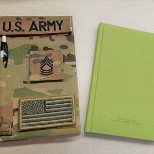 May include: A camouflage-patterned notebook cover with "U.S. ARMY" text, an American flag patch, and a pen holder. Next to it is a green notebook with the text "FEDERAL SUPPLY SERVICE".
