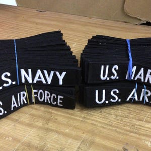 May include: Bundles of black fabric patches with white embroidered text. The patches read "U.S. NAVY", "U.S. AIR FORCE", "U.S. MARINES", and "U.S. ARMY". The patches are secured with blue and yellow rubber bands.