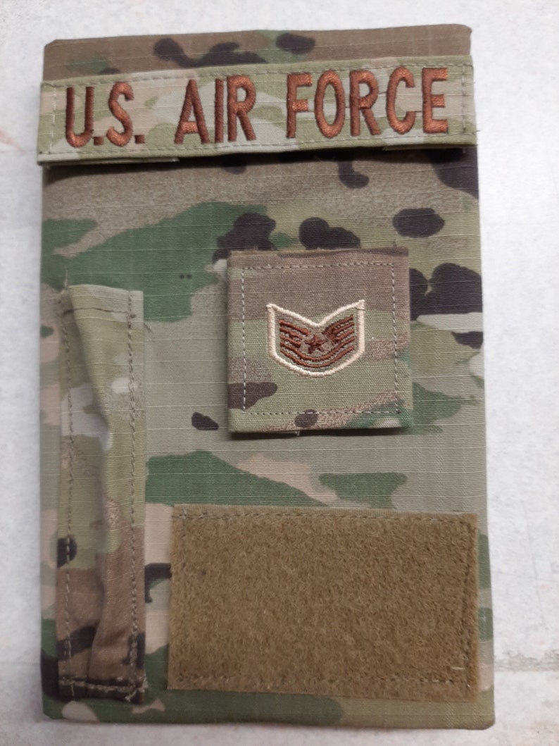 May include: Camouflage-patterned notebook cover with the text "U.S. AIR FORCE" embroidered in brown. Features a brown patch with a military rank insignia and a rectangular velcro patch.