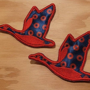 May include: Two red felt appliques of geese flying in formation. The geese are outlined in red and have a blue and red patterned fabric design.