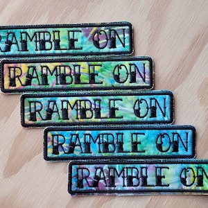 May include: Rectangular embroidered patches with the text "RAMBLE ON" in black block letters. The patches have a black border and a colorful, tie-dye-like background in shades of blue, green, yellow, and purple. The patches are arranged on a light wood surface.