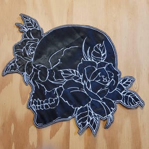 May include: A black fabric patch featuring a white embroidered outline of a human skull surrounded by two blooming roses with leaves. The design is rendered in a clean, minimalist line art style with high contrast between the black and white elements.