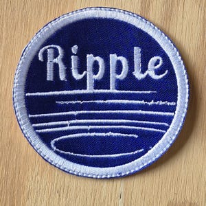 Könnte beinhalten: Ein blauer und weißer gestickter Aufnäher mit dem Wort "Ripple" in weißen Buchstaben. Der Aufnäher hat einen weißen Rand und ein Design aus weißen Linien, die an Wasserwellen erinnern.