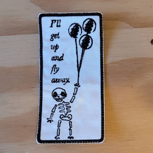 Peut inclure: Patch brodé blanc avec un squelette tenant trois ballons noirs. Le texte dit "Je vais me lever et m'envoler."