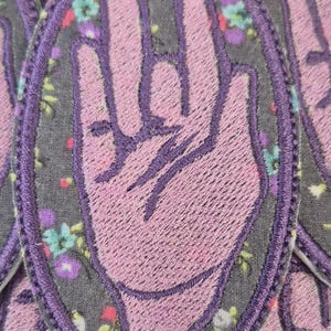 Puede incluir: Un parche bordado de forma ovalada que presenta un gesto de mano de color morado claro sobre un fondo gris oscuro con un pequeño estampado floral. La mano se muestra con el pulgar, el índice y el dedo medio extendidos, y los otros dedos doblados.