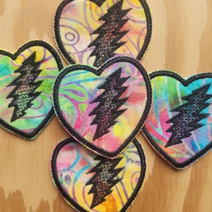 May include: Five colorful heart-shaped patches with a black lightning bolt design. The patches are made of fabric and have a tie-dye pattern.