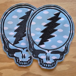 May include: Two embroidered patches featuring a skull design with a lightning bolt on a light blue background with white clouds. The patches have a black border and are placed on a wooden surface.