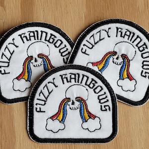 May include: Three white embroidered patches with black outlines. Each patch features a skull with a rainbow coming out of its eye sockets. The text "Fuzzy Rainbows" is written in a gothic font around the skull.
