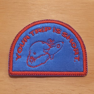 Pode incluir: Um remendo bordado azul e vermelho com o texto "Your Trip Is Short" e uma imagem de desenho animado de uma pessoa em um foguete.