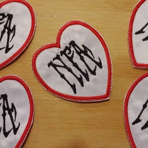 May include: Heart-shaped patches with a white satin finish, a red border, and the letters "NFA" in a gothic font. The patches are arranged on a wooden surface.