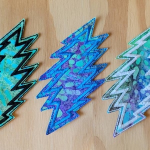 Könnte beinhalten: Drei gestickte Patches in Form eines Blitzes. Die Patches sind blau, lila und grün mit einem weißen Wirbelmuster. Der Umriss des Blitzes ist schwarz auf den blauen und grünen Patches und weiß auf dem lila Patch.
