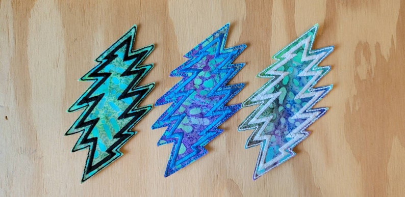Puede incluir: Tres parches bordados en forma de rayo. Los parches son azules, morados y verdes con un patr&oacute;n de remolino blanco. El contorno del rayo es negro en los parches azules y verdes y blanco en el parche morado.