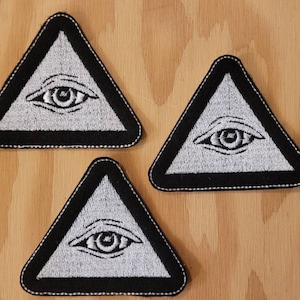 May include: Three black and white embroidered patches shaped like triangles with an eye inside each triangle. The eye is white with a black pupil.