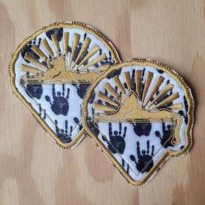 May include: Two white fabric patches with a black and white handprint pattern. Each patch features a gold embroidered sunburst design with a silhouette of a cat sitting on a crescent moon.