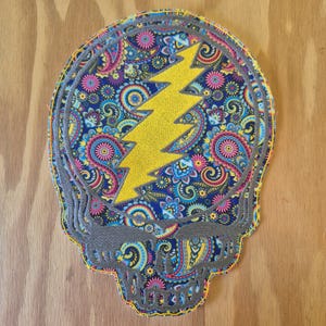 Può includere: Una toppa colorata con un teschio con un fulmine giallo. Il teschio è riempito con un motivo paisley nei toni del blu, rosa, giallo e verde, bordato di grigio.