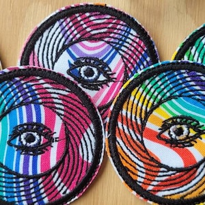 Könnte beinhalten: Fünf bunte gestickte Aufnäher mit einem wirbelnden Regenbogenmuster und einem schwarzen Auge in der Mitte. Die Aufnäher sind rund und haben einen schwarzen Rand.