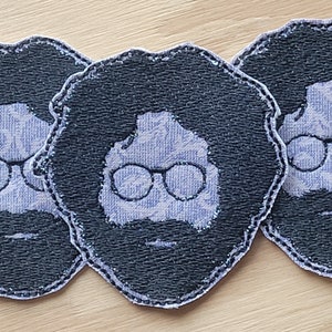 May include: Three black and white embroidered patches featuring a person with glasses and a beard. The patches are made of a textured fabric and have a stitched border.