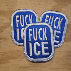 Może przedstawiać: Trzy niebieskie, haftowane naszywki z napisem "FUCK ICE" białymi literami. Każda naszywka ma białą obwódkę i zaokrąglony prostokątny kształt. Naszywki ułożone są na jasnobrązowej drewnianej powierzchni.