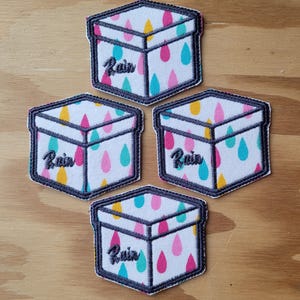 May include: Four white fabric patches shaped like boxes with a grey outline. Each patch has a colourful raindrop pattern and the word "Rain" written in black.
