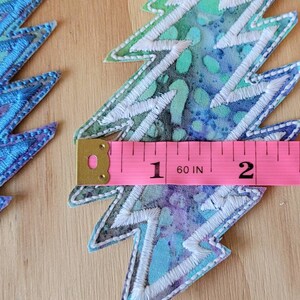 Puede incluir: Dos parches de rayo bordados en azul y blanco con una cinta m&eacute;trica rosa que muestra que tienen 2 pulgadas de ancho.