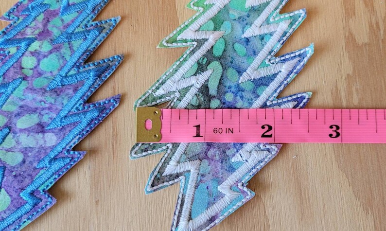 Puede incluir: Dos parches de rayo bordados en azul y blanco con una cinta m&eacute;trica rosa que muestra que tienen 2 pulgadas de ancho.