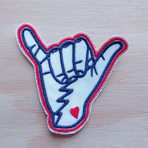 May include: White embroidered patch of a hand giving the shaka sign with a red heart tattoo on the wrist. The patch has a red and blue outline.