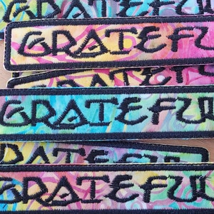 May include: A stack of colourful fabric patches with the word "GRATEFUL" embroidered in black on each patch. The patches have a tie-dye pattern and are edged with black stitching.
