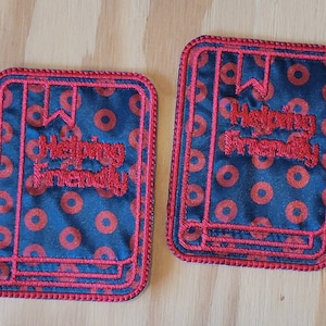 May include: Two red and blue fabric coasters with a book shape and the text "Helping is Friendly" embroidered on them.