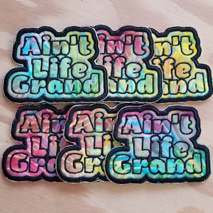 Peut inclure: Un ensemble de six patchs thermocollants colorés avec la phrase "Ain't Life Grand" dans un motif tie-dye. Les patchs sont parfaits pour ajouter une touche de personnalité aux vêtements, aux sacs ou à d'autres accessoires.