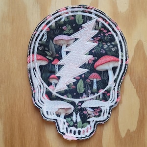 May include: A fabric patch in the shape of a skull with a pink lightning bolt in the center. The skull is filled with a dark floral pattern featuring red mushrooms and green leaves. The patch has a white border with a black outline.