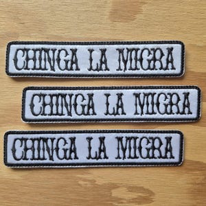 Puede incluir: Tres parches rectangulares con el texto "CHINGA LA MIGRA" bordado en negro sobre fondo blanco. Cada parche tiene un borde negro. Los parches están dispuestos sobre una superficie de madera.