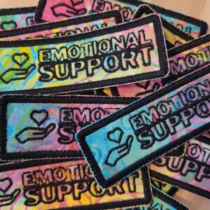 May include: A close-up of several colorful fabric patches with the text "Emotional Support" in black lettering. The patches have a tie-dye pattern and a hand with a heart in it.