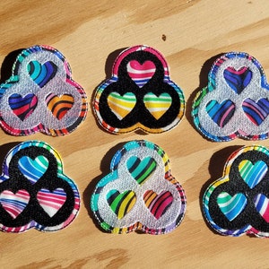 Puede incluir: Seis parches de tela coloridos con forma de trébol de tres hojas con tres corazones en el centro. Cada parche tiene una combinación de colores diferente de rayas y corazones.