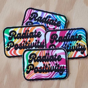 May include: Four rectangular patches with the words "Radiate Positivity" in black script. Each patch has a black border and a colorful, swirling rainbow design. The patches are on a light brown wooden surface.