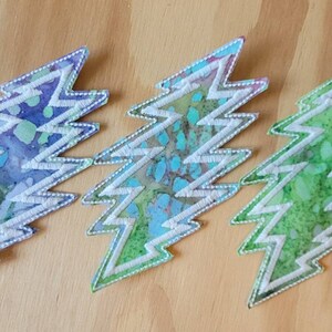Puede incluir: Tres parches bordados en forma de rayo. Los parches son blancos con un patr&oacute;n tie-dye azul, verde y morado.