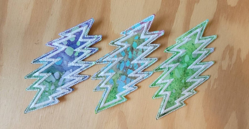 Puede incluir: Tres parches bordados en forma de rayo. Los parches son blancos con un patr&oacute;n tie-dye azul, verde y morado.