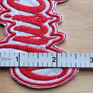 May include: Red embroidered patch with the word "S**" in a stylized font. The patch is approximately 2 inches wide and 1 inch tall.