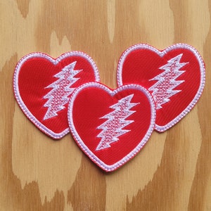 Op de afbeelding: Drie rode vilt hartvormige patches met witte bliksemontwerpen. De bliksem is wit omrand.