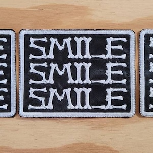 Op de afbeelding: Drie zwarte stoffen patches met witte geborduurde tekst die drie keer "SMILE" op elke patch zegt.