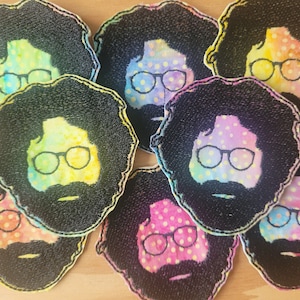 Könnte beinhalten: Ein Set aus acht gestickten Aufnähern mit einem Cartoon-Gesicht mit buschigem Afro, Brille und Bart. Die Aufnäher sind bunt und haben einen Batik-Effekt mit weißen Punkten.