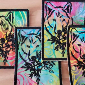 Puede incluir: Conjunto de cuatro parches de tela rectangulares con un fondo tie-dye colorido. Cada parche presenta una cabeza de lobo bordada en negro y un diseño floral. Los parches tienen un borde negro y están dispuestos sobre una superficie de madera.