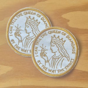 May include: Two white embroidered patches with gold trim and text. Each patch features a profile of a queen wearing a crown, holding a diamond. The text reads "I CAN TELL THE QUEEN OF DIAMONDS BY THE WAY SHE SHINES".
