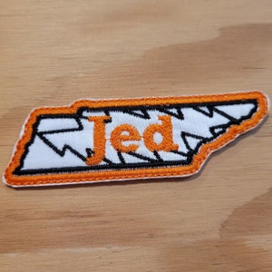 Op de afbeelding: Een oranje en witte geborduurde patch in de vorm van de staat Tennessee met het woord "Jed" in oranje letters.