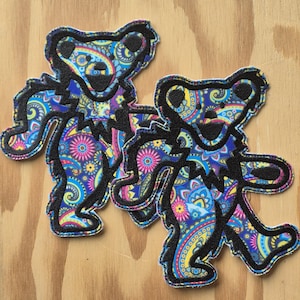 Peut inclure: Deux écussons en forme d'ours avec un motif cachemire coloré en bleu, jaune, rose et violet. Les ours ont des contours noirs et sont posés sur une surface en bois brun clair. Idéal pour les vêtements ou accessoires.